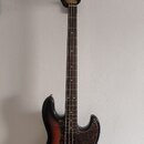 Elektrická basová kytara s tělem v provedení sunburst a hnědým hracím hřidlem. Tělo má tmavý vzorovaný pickguard a na hlavě je viditelné logo Fender.…