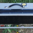 Šedý transportní kufr s černými kovovými rohy a černé rukojetí. Na horní straně je umístěna černá ventilační mřížka a kulatý žlutý štítek. Zadní stra…