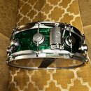 Zelený snare drum s kovovými obručmi a mechanismem na straně. Na mechanismu je viditelné logo dbs. Povrch nástroje vykazuje známky opotřebení v podob…