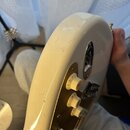 Detail zadní strany bílé elektrické kytary s kovovým zlatým krytem elektroniky. Na kytaru jsou vidět početné škrábance a drobné oděrky laku. Jsou zde…