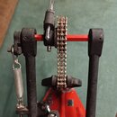 Černý bicí pedál s červenými kovovými prvky. Na mechanismu je vidět pružina a řetězový pohon. Povrch černých částí působí matně a je mírně znečištěný…