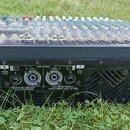 Černý mixážní pult Yamaha POWERED MIXER MODEL EMK50VNC položený na trávě. Na zadním panelu jsou viditelné konektory pro reproduktory SPEAKER S a A, n…