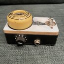 Kytarový efekt v černé krabičce s bílým horním panelem. Na vrchu je umístěno žluté kolečko s nápisem WHEEL GUITAR PEDAL SERIES a logem ve tvaru písme…