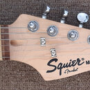 Fotografie zachycuje hlavu a část hmatníku elektrické kytary z přírodního světlého dřeva. Na hlavě je černý nápis Squier MINI by Fender®. Jsou vidět…