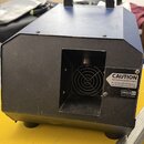 Modrý a černý přístroj s hranatým tvarem, který má v přední části mřížku ventilátoru. Na pravé straně je bílá nálepka s nápisem CAUTION Hot Surface!…