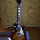 Elektrická kytara s tónem sunburst, bílým pickguardem a dvěma humbuckery. Na hlavě je nápis BaCha a pod ním logo ve tvaru stylizovaného písmena V. Hр…