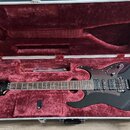 Černá elektrická kytara v hliníkovém kufru s červeným plyšovým vnitřkem. Na hlavě nástroje je bílý nápis Ibanez. Kytara má dva humbuckery, tremolo mo…