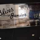 Kovový štítek s nápisem Blues Junior. Je na něm uveden text A PRODUCT OF: FENDER MUSICAL INSTRUMENTS CORP.. a sériové číslo B-153256. Štítek vykazuje…
