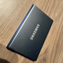Na fotografii je modrý obdélníkový předmět s kovovým povrchem, který leží na dřevěném stole. Uprostřed je bílý nápis SAMSUNG. Povrch předmětu vypadá…