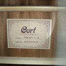 Fotografie detailu vnitřní štítku akustické kytary. Na štítku je logo Cort GUITARS, označení MODEL: EARTH70 W NS a SERIAL NO: CA211007408. Dole je uv…