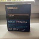 Fotografie zobrazuje modré papírové balení s produktem značky SHURE. Na krabici jsou nápisy LEGENDARY PERFORMANCE™, GLX-D WIRELESS a model SB902A. Te…