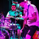 Fotografie zachicuje kapelu během koncertu s bubeníkem a kytaristou. Bubeník hraje na sadbu s činely značky PAISTE a bubny, které mají bílý nápis the…