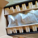 V otevřené kartonové krabici je předmět zabalený v bílé plastové fólii. Fixaci zajišťují dvě formované vložky z recyklovaného papíru. Na fólii je vid…