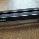 Černý obdélníkový podstavec s dvěma paralelními protiskluzovými pásky z tmavého materiálu. Na přední straně je bílý nápis D'Addario. Nástroj stojí na…