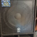 Černý basový reproduktor v bedně potažené tmavou látkou s kovovou mřížkou. V horní části je modrá nálepka s nápisy YAMAHA, AX SERIES, Great Sounds •…
