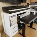 Černý digitální klavír umístěný na bílém stole s přihrádkami. Na levé straně klavíru je nápis MONTREUX. K sestavě patří černá polstrovaná piano stoli…