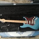 Modrá elektrická kytara v černém tvrdém kufru. Na hlavě hředu je viditelné logo Fender a v horní části kufru nápis Fender. Kytara má javorový hrideln…