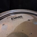 Fotografie detailu průhledné bubny na bicí nástroj. Na povrchu je černý nápis EVANS a pod ním označení EC2S. Povrch vykazuje drobné škrábance a nečis…