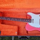 Růžová elektrická kytara v černém kufru s oranžovou výplní. Na hlavě hředu je logo Fender. Kytara má bílý pickguard a kovové části s viditelnými stop…