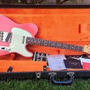 Růžová elektrická kytara s bílým pickguardem v otevřeném černém kufru s oranžovou výstelkou. Na hlavě kytary je viditelné logo Fender. V kufru jsou v…