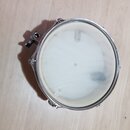 Kruhlový bicí buben s bílým průsvitným blánou a chromovaným obručem. Na levé straně je vidět kovový mechanismus pro uchycení. Povrch blány vykazuje m…