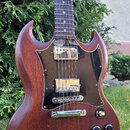 Elektrická kytara s hnědým dřevěným povrchem a tmavým hmatníkem. Má černý plecháč, dva humbuckery s chromovanými kryty a čtyři černé ovládací knoflík…