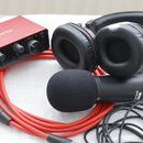 Sada audio vybavení tvořená červeným zvukovým rozhraním, černými sluchátky a černým mikrofonem s černé molítkem. Na rozhraní je bílý nápis Focusrite…