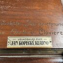 Detail dřevěného povrchu klavíru s vyrytým nápisem „Höchste Auszeichnung Für Oesterr. Claviere“. Pod ním je přilepený kovový štítek s textem „VELKOSK…