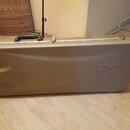 Zlatý tvrdý kufr na hudební nástroj s černou rukojetí a kovovými zapínacími zámky. Na pravé straně je vyražen nápis Gibson. Povrch kufrů vykazuje dro…