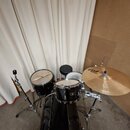 Černá bicí souprava s kovovým hardwarem, zahrnující basový buben, dva tom-tomy a malý buben. Na činely je viditelné logo "Paiste". Souprava obsahuje…