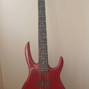 Čtyřstrunná elektrická basová kytara s červeným trupem a tmavým hmatníkem. Na hlavě nástroje je viditelné logo ve tvaru písmena H. Trup vykazuje znám…