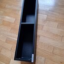 Černý úhelkový stojan s dvěma přihrádkami, umístěný na dřevěné podlaze. Na spodní hraně je vidět bílá nálepka s nápisem GLORIOUS. Povrch je matný a v…