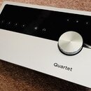 Stříbrný audio rozhraní s černým ovládacím panelem a velkým kovovým otočným knoflíkem. Na šasi je vlevo logo a nápis APOGEE a vpravo nápis Quartet. N…
