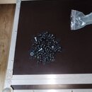Fotografie zachycuje otevřený transportní kufr s hnědým vnitřním dnem a hliníkovými profily. Uprostřed dna je rozsypaná hromadka černých šroubů s pod…