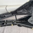 Černá elektrická kytara s tělem ve tvaru V, položená v otevřeném černém tvrdém kufru s šedým vnitřkem. Na hlavě nástroje je bílý nápis Gibson. Kytara…