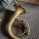 Zlatý mosazný barytonsaxofon položený na koberci. Na zvonu je viditelné kulaté logo, které je však nečitelné. Povrch nástroje vykazuje známky opotřeb…