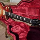 Elektrická kytara v barvě sunburst s tmavě červeným okrajem, položená v otevřeném hnědém kufru s červeným vnitřkem. Na hlavě kytary je bílý nápis „Le…