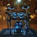 Elektronická bicí souprava značky Roland v černé barvě s modrým podsvícením. Na basovém bubnu je bílý nápis Roland a V-Drums, na činelech je viditeln…