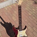 Elektrická kytara s červeným lakovaným tělem a hnědým hmatníkem. Na hlavě je viditelné logo Squier. Kytara má bílý pickguard a tři snímače. Nástroj s…