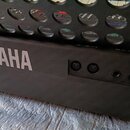 Detailní záběr na zadní část černého klavíru s bílým nápisem YAMAHA. Jsou vidět ovládací prvky v podobě otočných knoflíků a několik připojovacích kon…