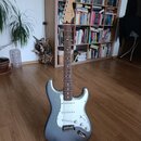 Stříbrná elektrická kytara s bílým pickguardem a javorovým hnedým hmatníkem. Na hlavě je viditelné logo Fender. Kytara stojí na černém stojanu v inte…