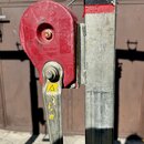 Na fotografii je červený kovový mechanismus s klikou připevněný k hranatému kovovému sloupu. Na červeném krytu jsou viditelné varovné piktogramy v tr…