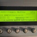 Detailní záběr na displej a ovládací prvky elektronického hudebního nástroje. Zelený LCD displej zobrazuje text: „A002:Square Hollow“, „Synth-Soft“,…