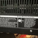 Detailní záběr na zadní část černého elektronického zařízení s ventilační mřížkou. Jsou zde dvě bílé nálepky s nápisy Tube Status Monitor a Tube Matc…