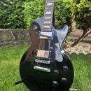 Černá elektrická kytara s tvarem typu Les Paul a hnědým hmatníkem. Na kytaru jsou instalovány dva humbuckery s kovovými kryty a čtyři ovládací knoflí…