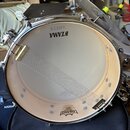 Fotografie zachycuje snare drum s kovovým obručím a bílým blátcem. Na blátci jsou viditelné nápisy TAMA AMATH a REMO. Přes bláto je natažený kovový p…