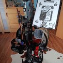 Sestava hudebního vybavení na černém stojanu. Je zde vidět kytara v červeném lakovaném provedení, mikrofon s šedým chlupatým krytem a tuner značky JO…