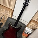 Elektrická kytara s tmavě červeným gradientem (sunburst) a černým hmatníkem. Na hlavě nástroje je viditelné logo ESP. Kytara leží na šedém křesle ved…