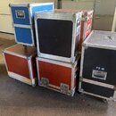 Skupina pěti transportních case různých velikostí a barev (modrá, červená, černá) s kovovými hranami a rohy. Na jednom z černých case je bílý nápis P…