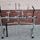 Kovový stojan na bicí bubny s několika nastavitelnými rameny a úchopy. Konstrukce je stříbrná, s černými plastovými knoflíky a koncovkami nohou. Na s…